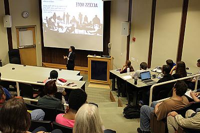 礼堂后面的照片显示的是Lydia X.Z. Brown addressing the audience. 他们穿着深色的夹克，打着领带. 在他们身后是一个屏幕投影，上面写着“访问笔记”，并包含几个剪影人物